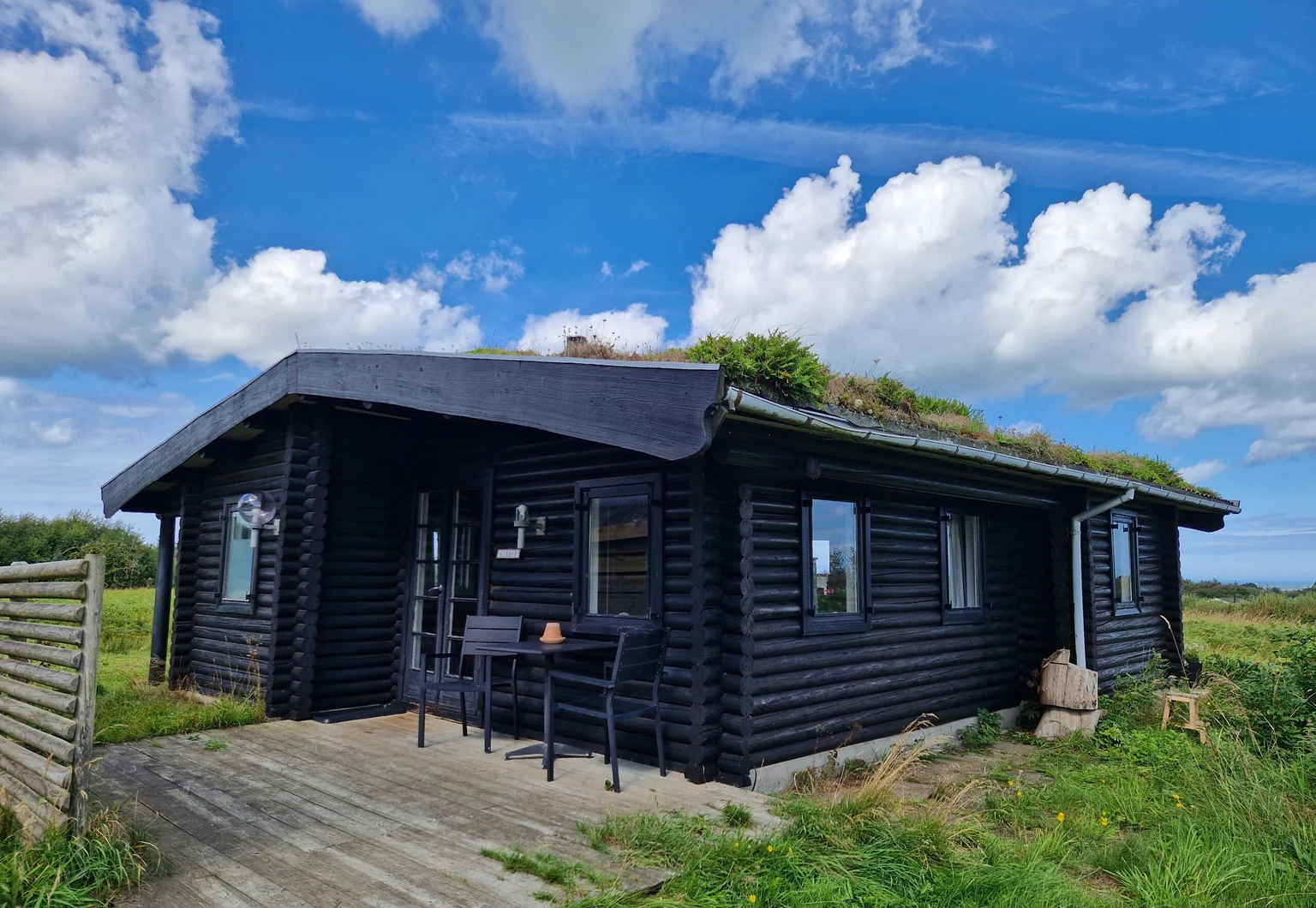 Das schwarze Blockhaus mit Gründach und Holzterrasse unter blauem Himmel mit Wolken